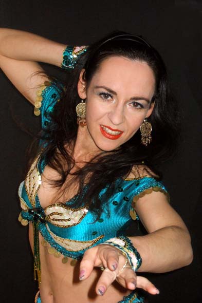 Tancerka brzucha w turkusowym kostiumie z frędzlami, złotymi kolczykami i ciemnymi włosami, pozująca na czarnym tle z wyciągniętą dłonią.